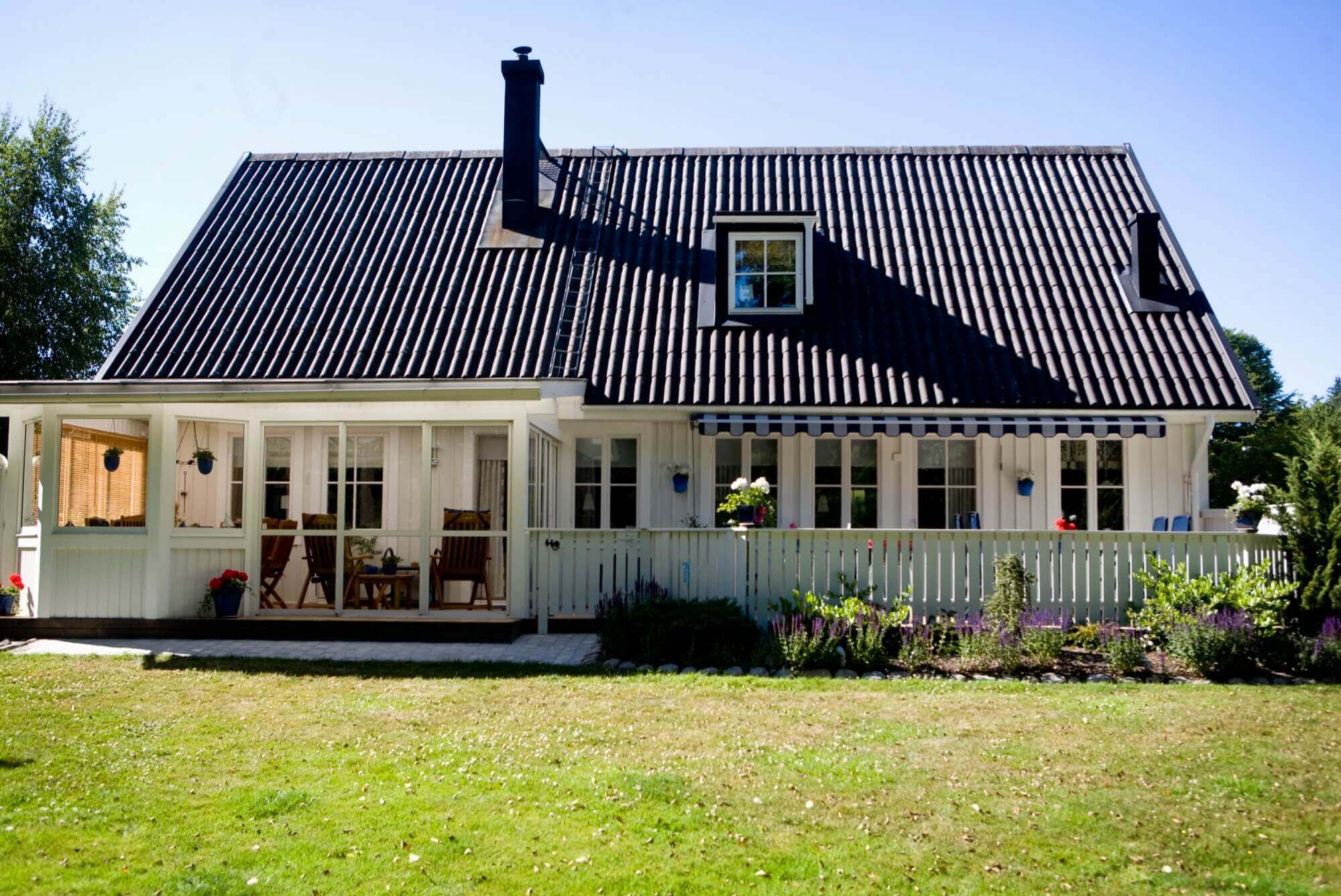 Valkoiseksi maalattu puuhuvila, jossa on suuri kuisti, musta katto ja vehreä puutarha - hyvin hoidettu julkisivumaalaus.